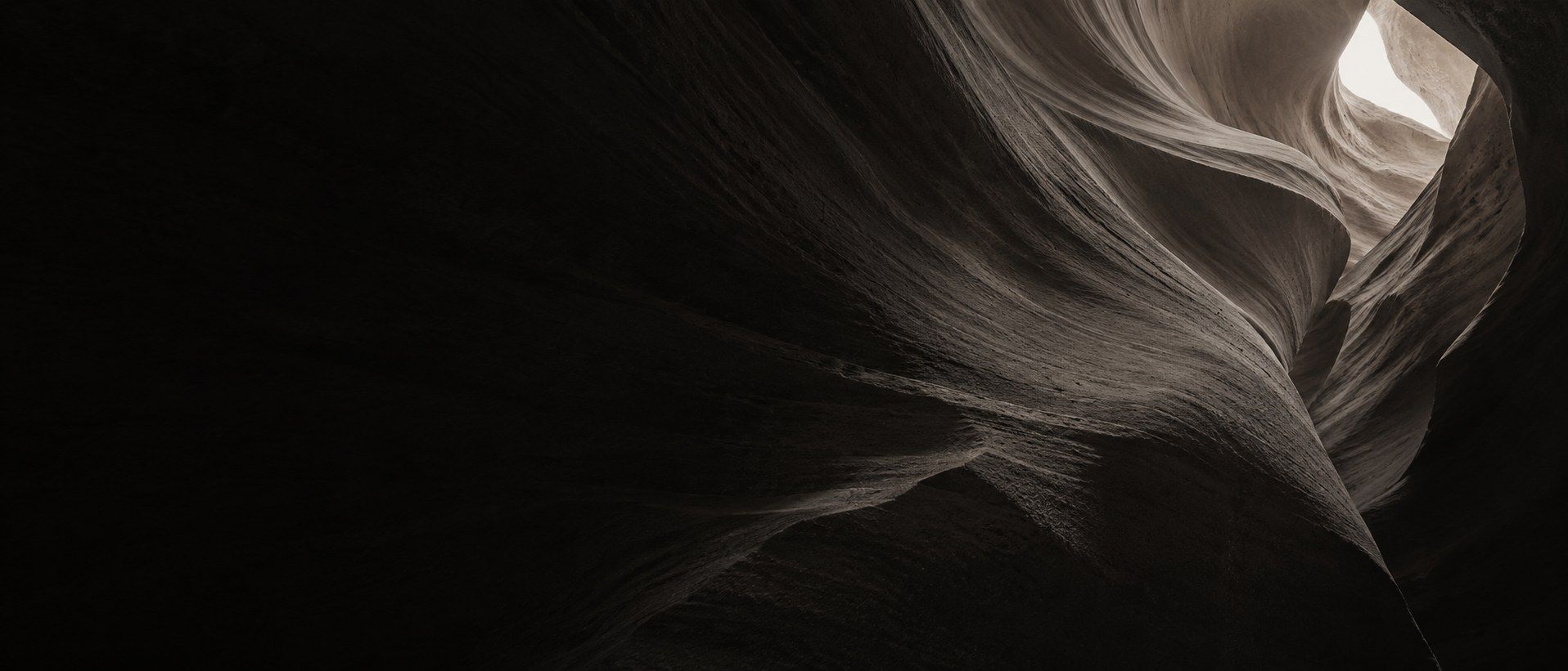 Wind-worn canyon rock in flowing dark layers, with pale light at an opening in the upper right.