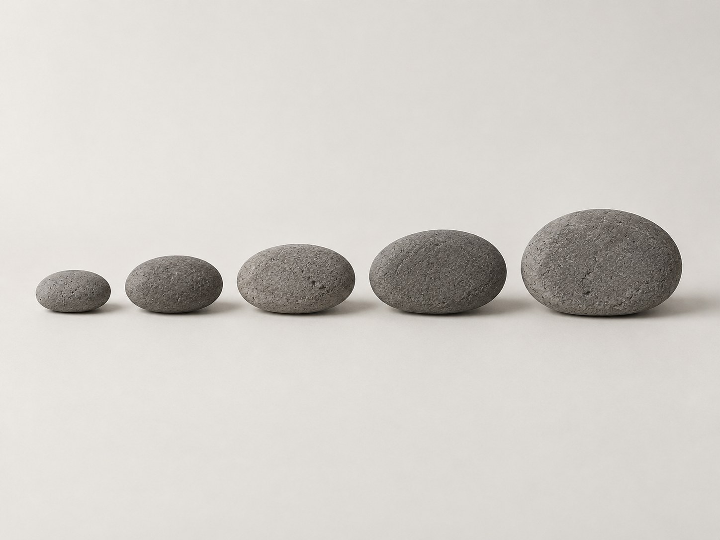 Five smooth grey stones in a line on a warm white ground, growing from small at the left to large at the right.