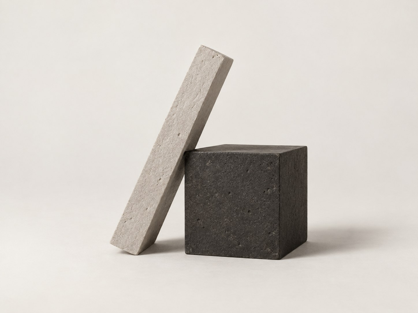 A tall thin pale stone slab leaning against a dark stone cube on a warm white ground.